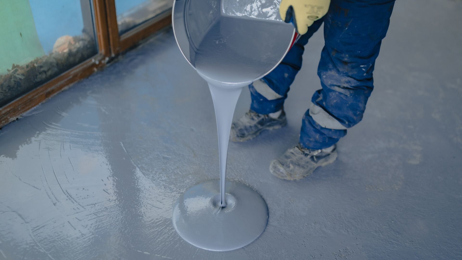 Worker pouring gray liquid floor coating from bucket onto concrete surface