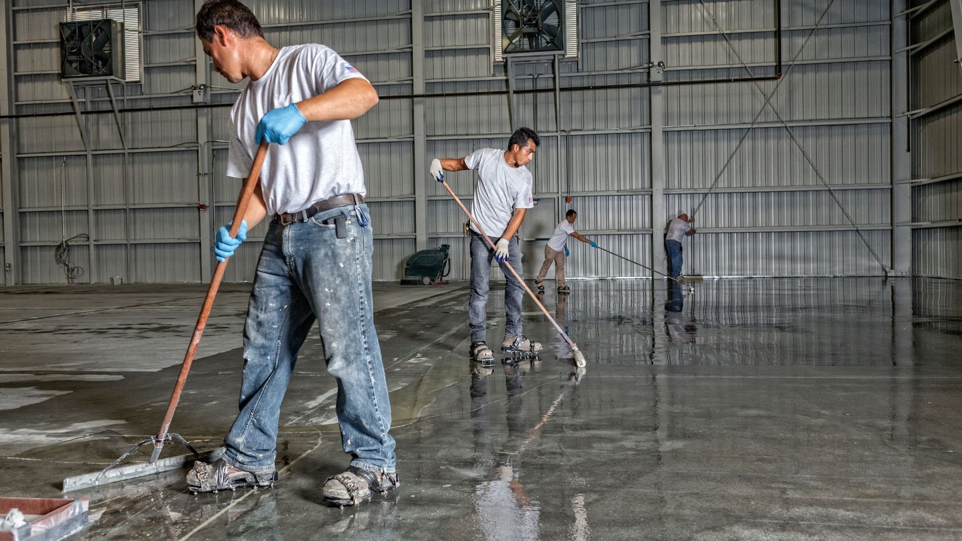 Workers mopping and cleaning large industrial warehouse floor with long mops